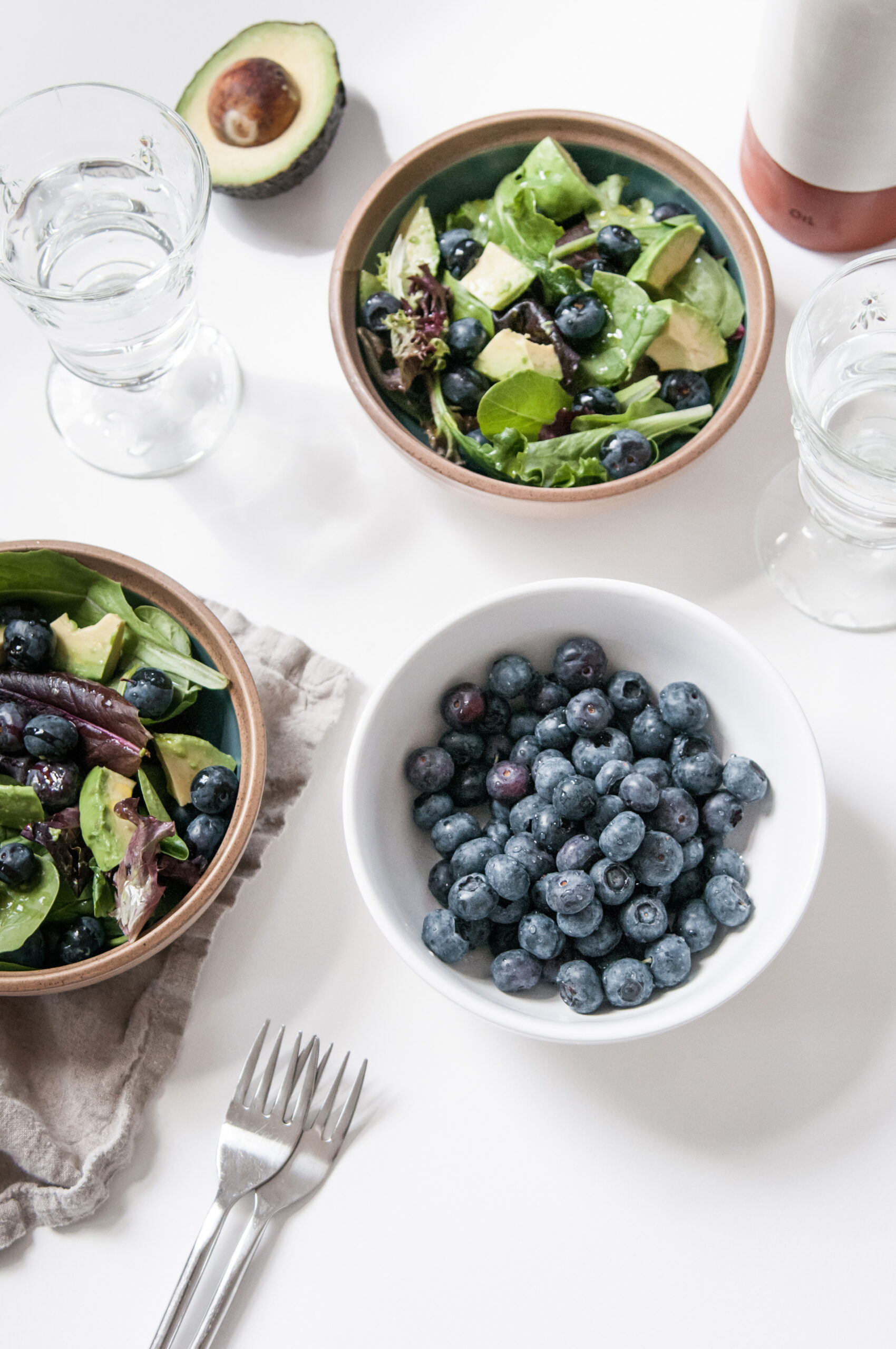 A fresh and healthy meal featuring bowls of blueberry avocado salad, a bowl of blueberries, glasses of water, and a halved avocado on a white table.