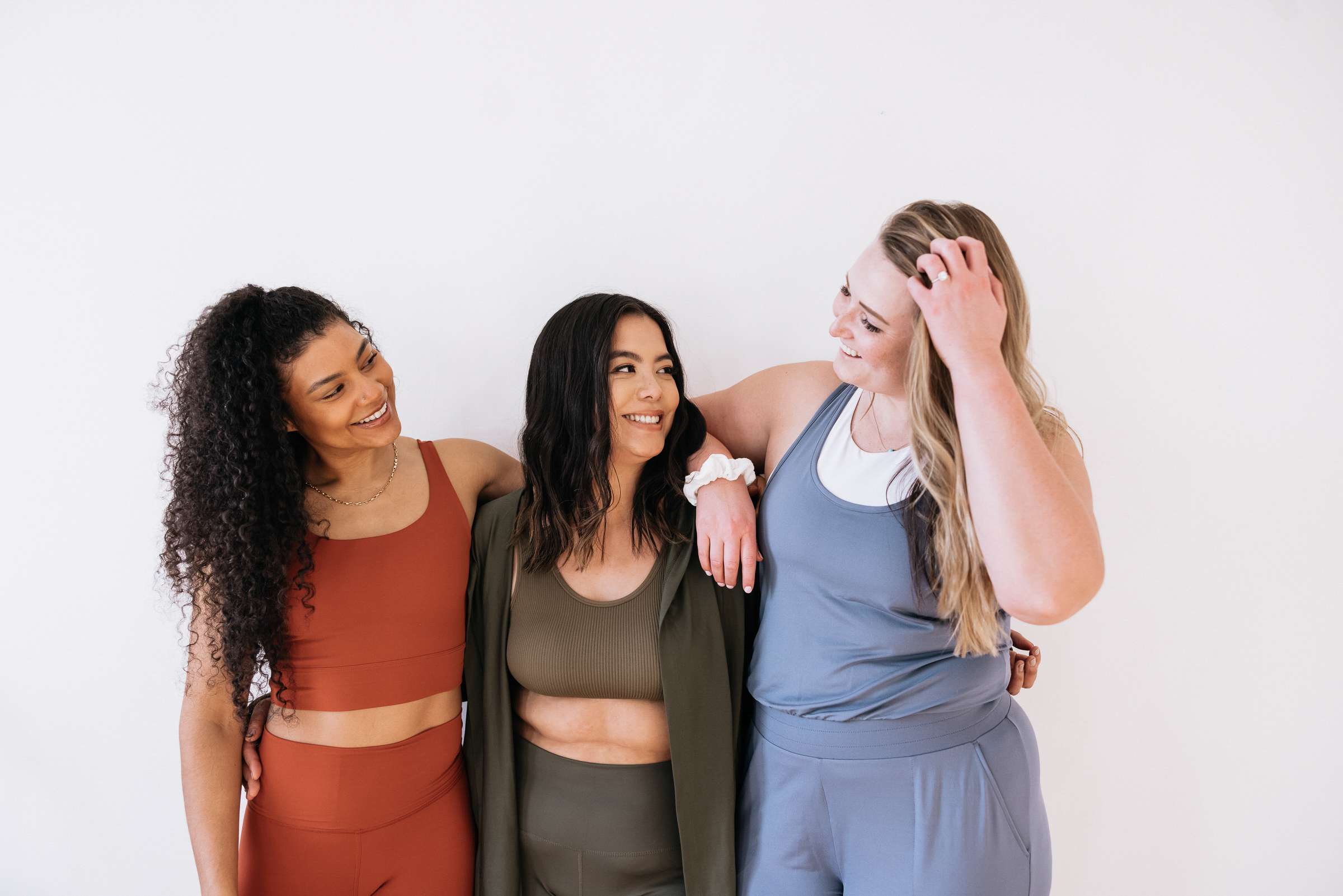 Three women in stylish activewear smile and embrace each other against a white background, showcasing body positivity and friendship.